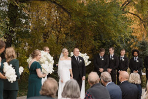 A bride and groom stand together outdoors during a wedding ceremony. Bridesmaids in green dresses and groomsmen in black suits are nearby, surrounded by autumn foliage and guests seated in front.