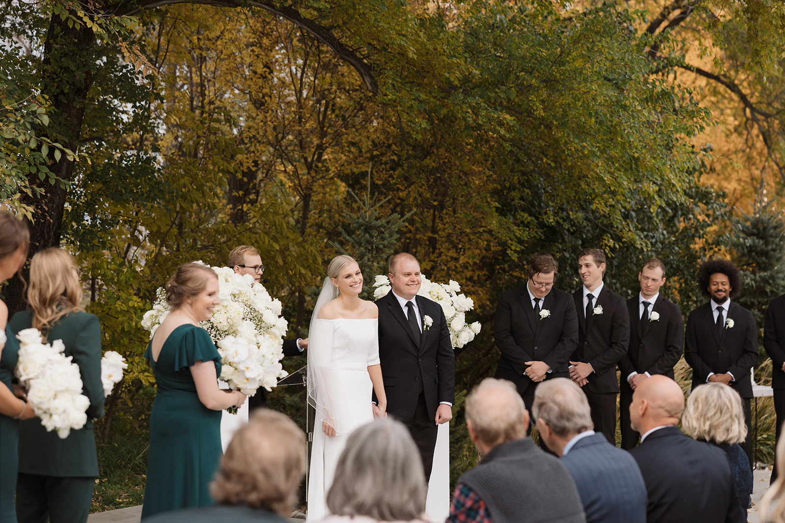 A bride and groom stand together outdoors during a wedding ceremony. Bridesmaids in green dresses and groomsmen in black suits are nearby, surrounded by autumn foliage and guests seated in front.