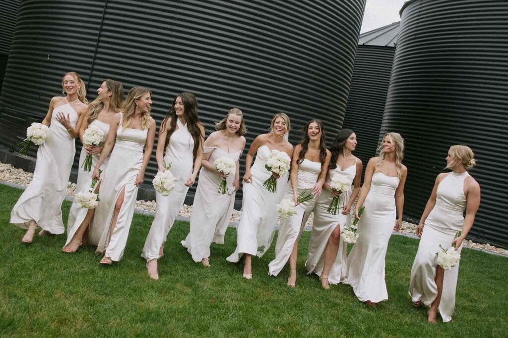 Bridal party walking by the black silos at The Pines, with bridesmaids in white dresses holding matching bouquets for a chic and modern wedding look.