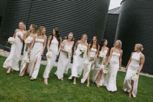Bridal party walking by the black silos at The Pines, with bridesmaids in white dresses holding matching bouquets for a chic and modern wedding look.