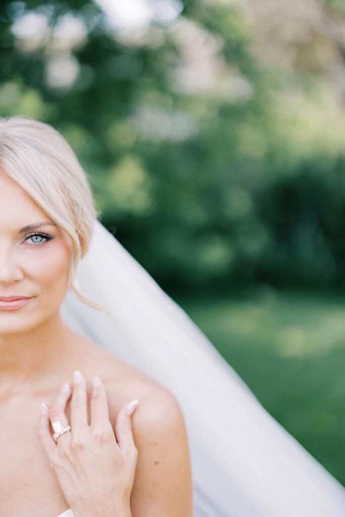 Close-up portrait of a bride, capturing her radiant smile, flawless makeup, and delicate veil, with soft greenery in the background at The Pines.