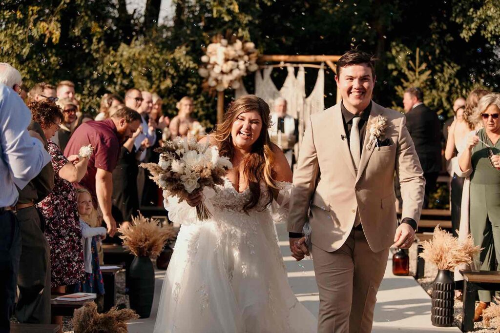 Joyful bride and groom exiting their wedding ceremony with dried bouquet in hand, greeted by smiling guests.