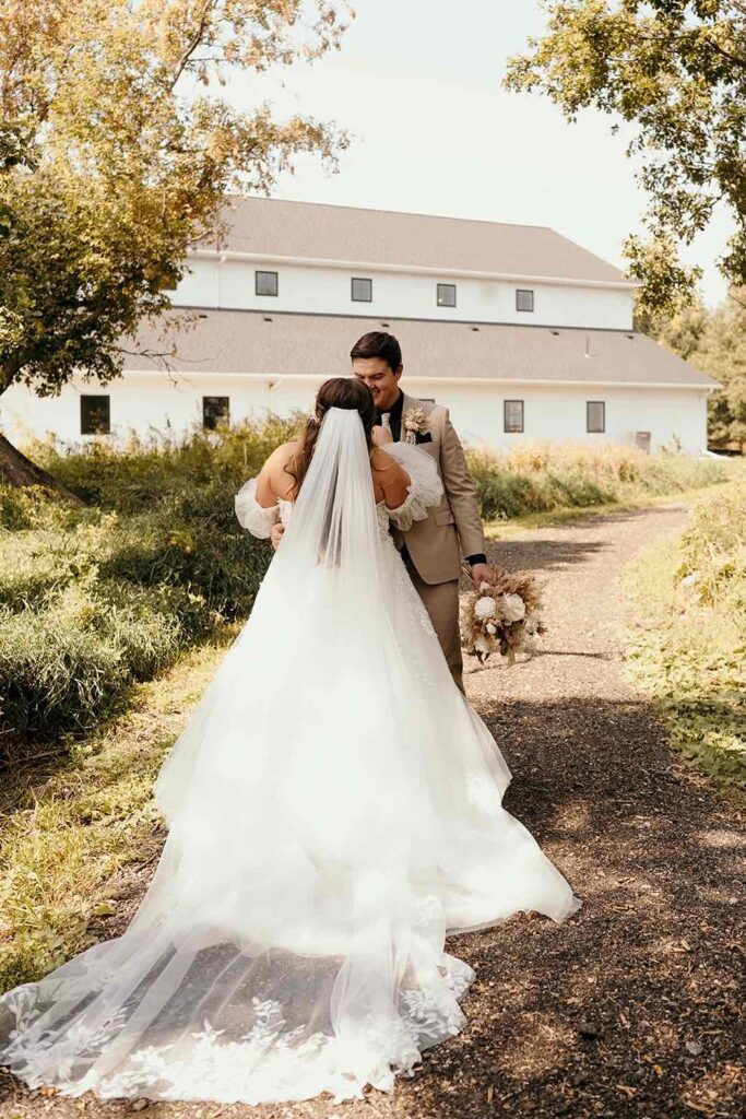 Rustic wedding portrait of bride and groom standing near a countryside barn surrounded by natural greenery.