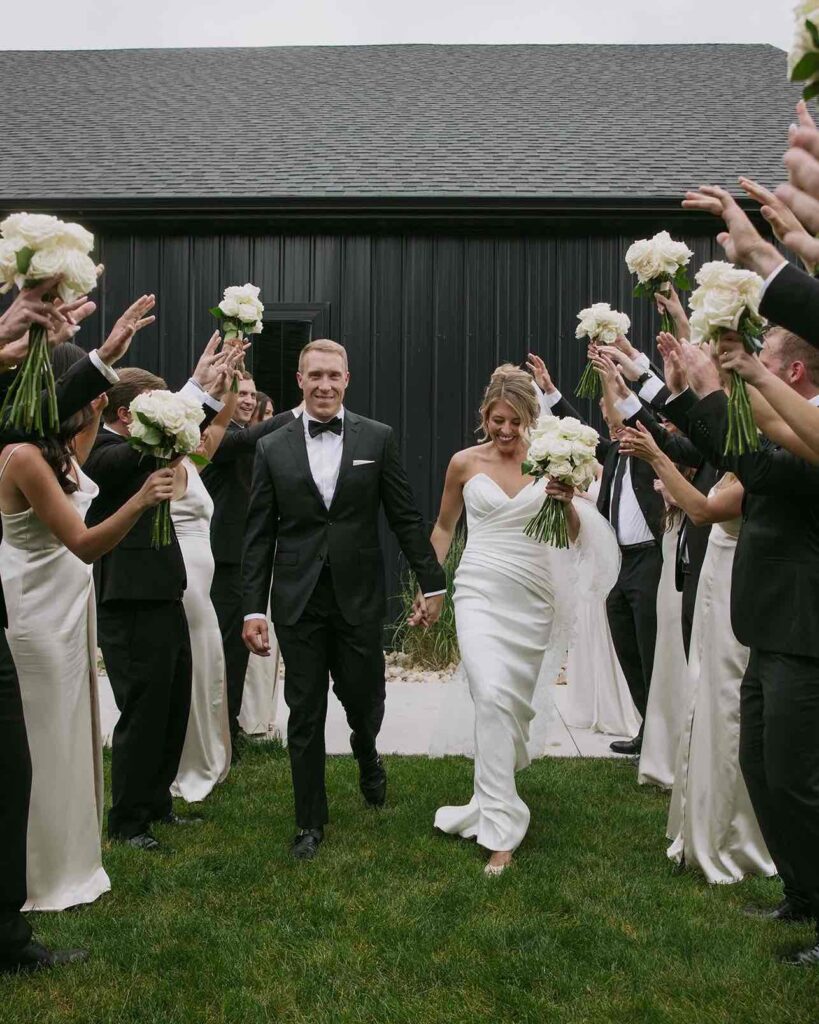 Bride and groom exiting ceremony at The Pines, walking hand in hand through a flower tunnel created by the wedding party, set against the black venue.