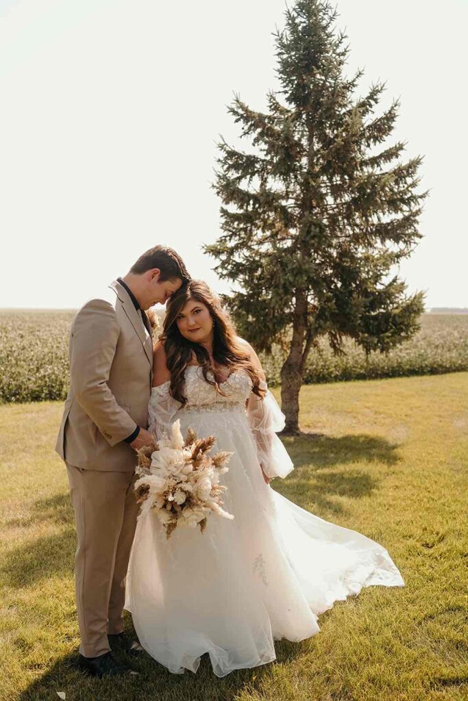 Bride and groom posing in a serene outdoor setting with the bride holding a dried floral bouquet and a single pine tree in the background.