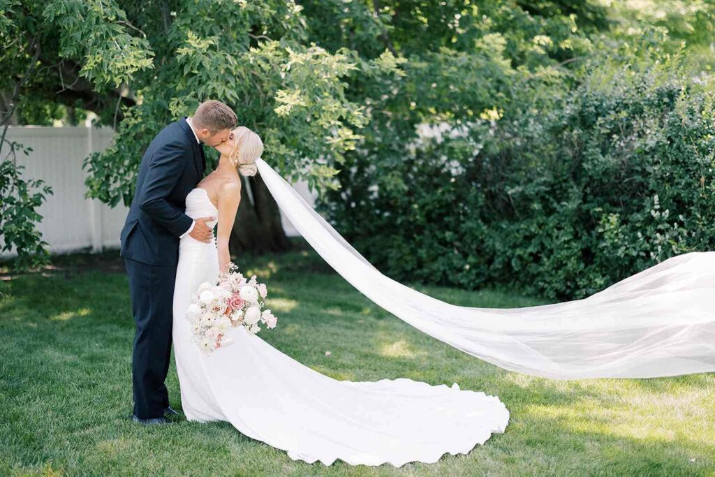 Bride and groom sharing a romantic kiss, the bride’s dramatic veil flowing behind them in a scenic outdoor setting at The Pines.