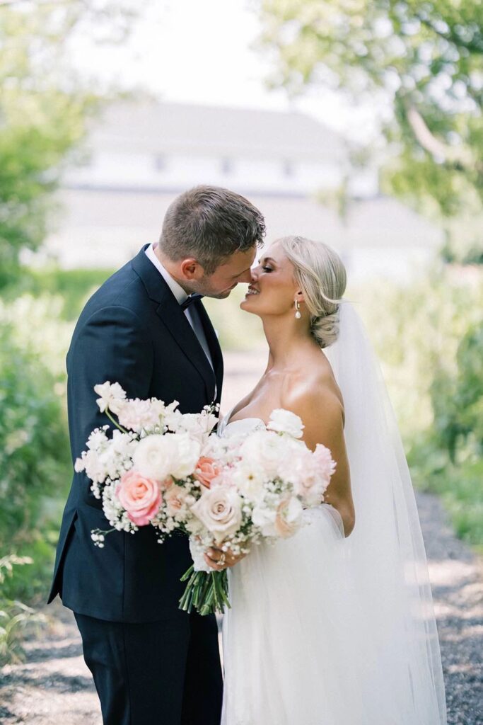 Bride and groom sharing a tender moment outdoors at The Pines, holding a beautiful floral bouquet with soft pink roses and white flowers, as they smile at each other.