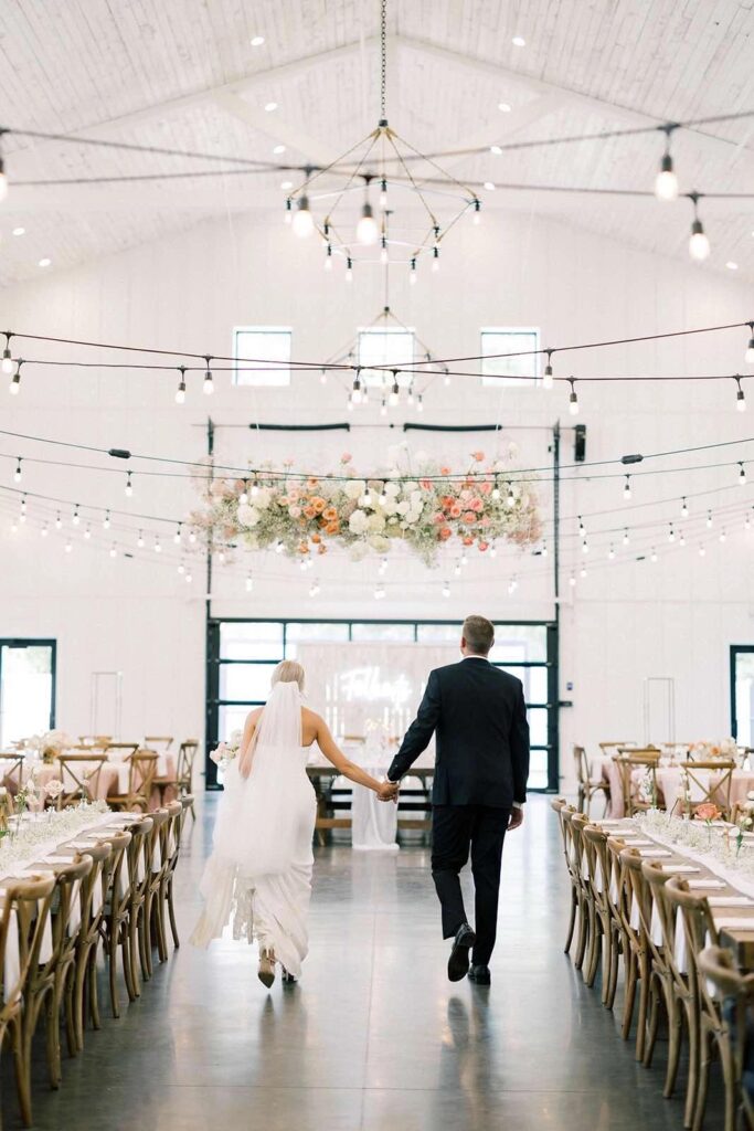 Bride and groom walking through the reception space at The Pines White Barn, with romantic floral arrangements overhead.