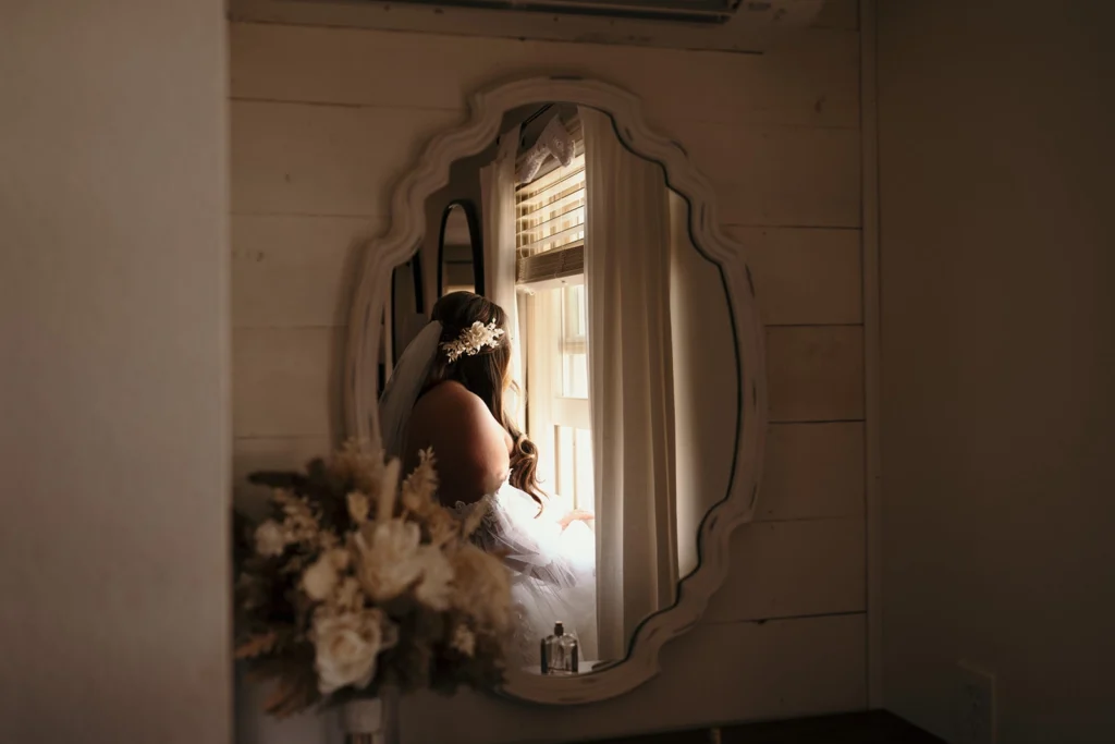 A bride, adorned with a floral hair accessory and veil, gazes out of a window. Her reflection is seen in an ornate, vintage-style mirror, with soft light illuminating the rustic bridal suite.