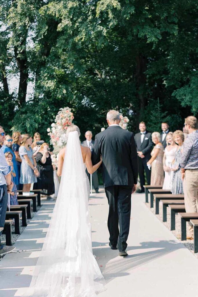 Bride walking down the aisle with her father at The Pines, surrounded by smiling guests and a breathtaking floral wedding arch.