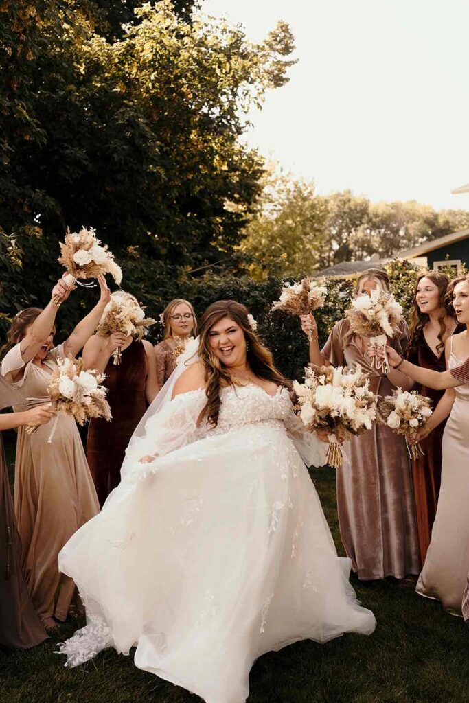 A joyful bride in a lace wedding gown surrounded by bridesmaids wearing neutral-toned dresses. Each bridesmaid holds a dried floral bouquet, celebrating outdoors in a lush green garden.