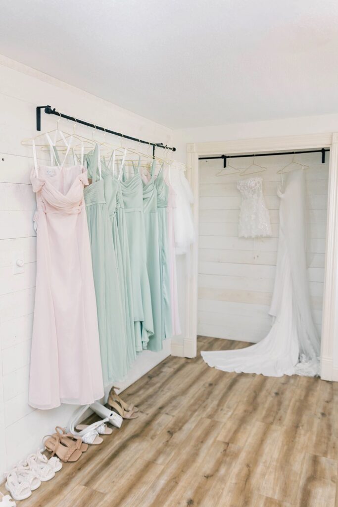 A row of pastel bridesmaid dresses and the bride’s wedding gown hang elegantly in the getting-ready suite at The Pines.