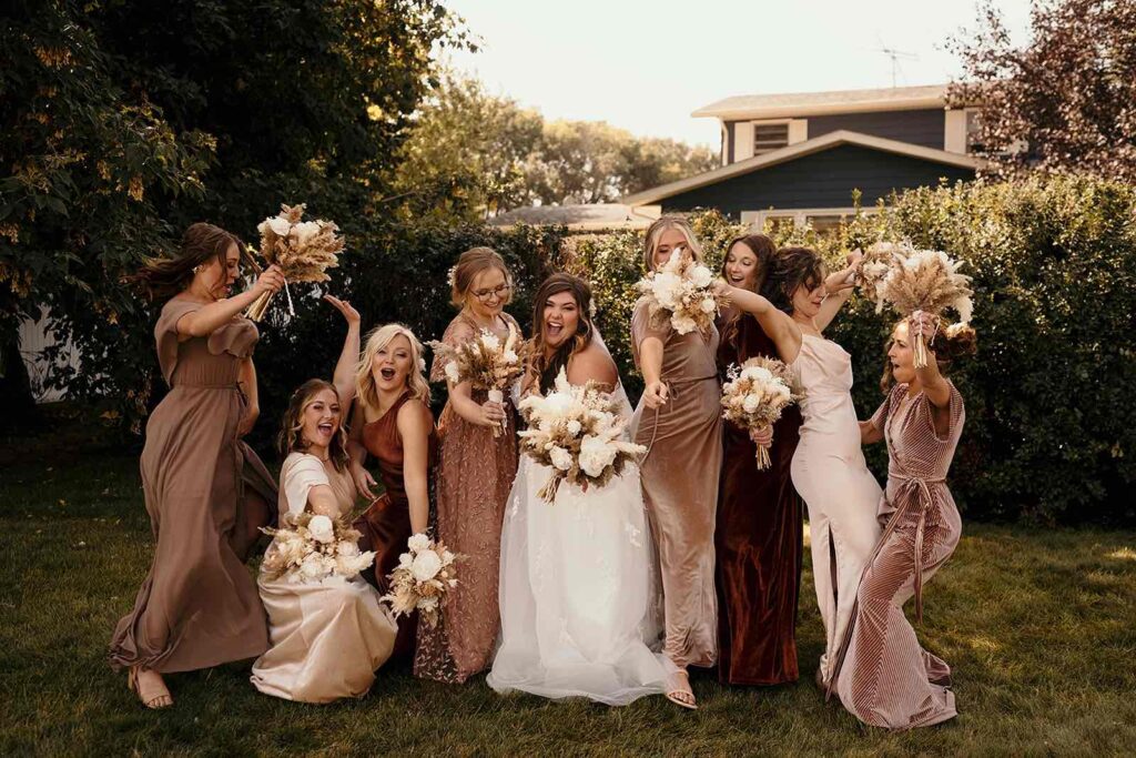 A lively group of bridesmaids in earthy-toned dresses, holding dried bouquets and celebrating with the bride, who is laughing and twirling in her wedding gown.