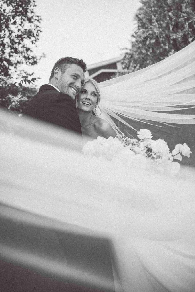 Black and white cinematic shot of the bride and groom smiling, with the bride’s flowing veil creating a dreamy, romantic wedding moment.