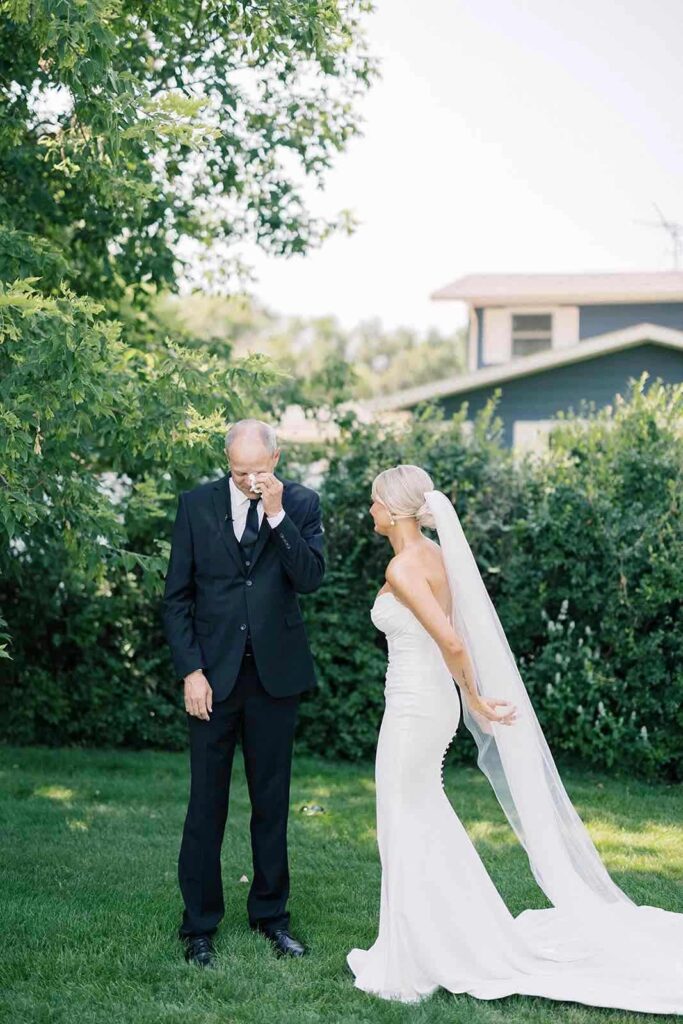 A bride, arm-in-arm with her father, walks down the outdoor aisle at The Pines as guests look on, surrounded by lush greenery.