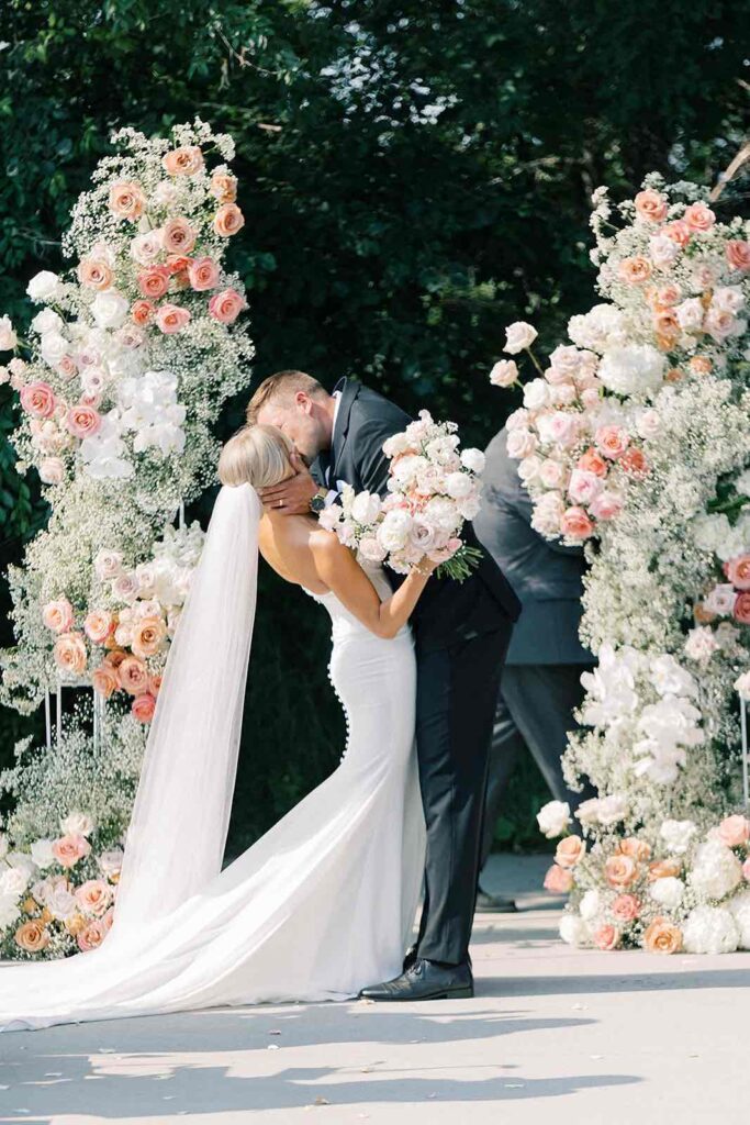 Newlyweds share a kiss while holding a pastel floral bouquet, as their wedding party moves around them in a dreamy, blurred effect.