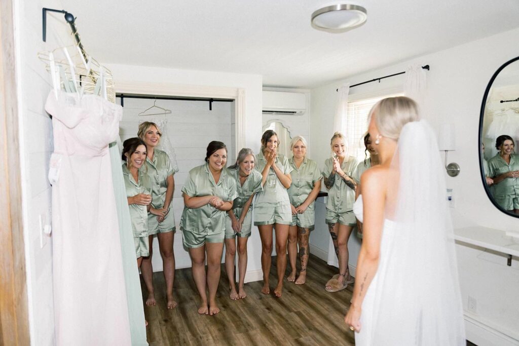 Bridesmaids in matching sage satin pajamas react with excitement as the bride reveals her wedding gown in The Pines' bridal suite.