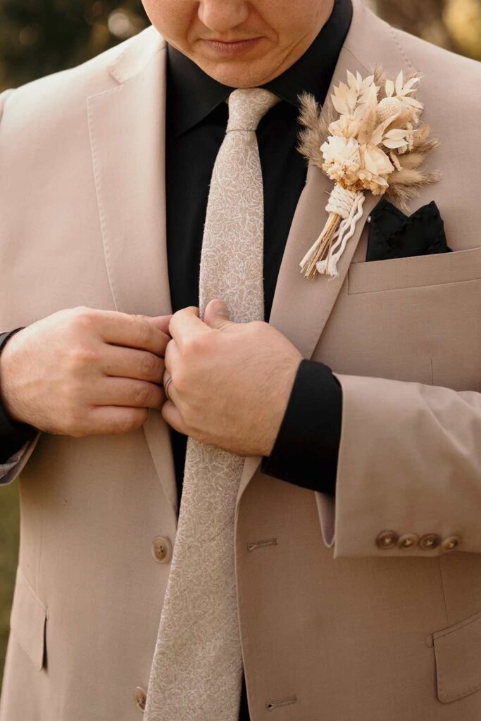 A close-up of a groom in a tan suit adjusting his tie, wearing a dried floral boutonniere with natural, bohemian tones.