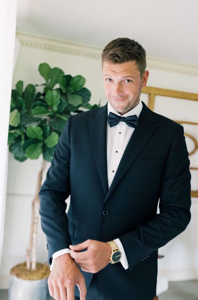 A groom in a classic black tuxedo adjusts his cufflinks in a stylish groom’s suite at The Pines.
