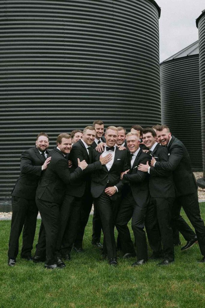Fun and candid group photo of groomsmen in black tuxedos at The Pines, posing by black silos for a modern wedding portrait.