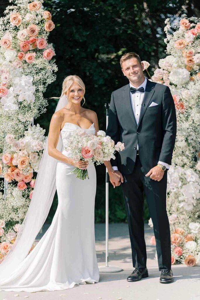 Just married couple standing by the ceremony floral arch at The Pines, beaming with joy.