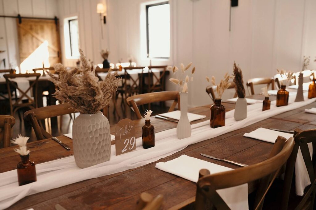 A long wooden table styled with neutral-colored vases, dried floral arrangements, amber glass bottles, and minimalist table settings, creating a rustic yet elegant ambiance.