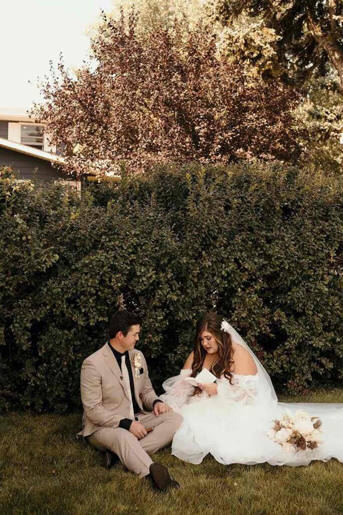 A bride and groom sit together on the grass, with the bride reading her vows. The backdrop features lush greenery, and the bride’s bouquet of dried florals rests nearby.