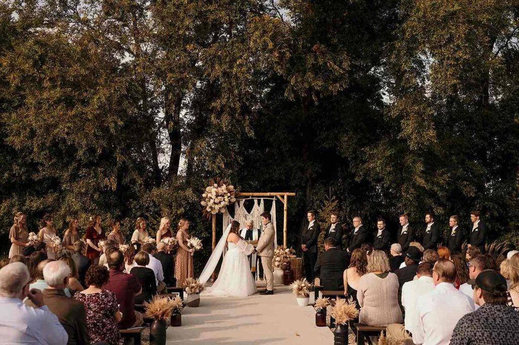 An outdoor wedding ceremony featuring the bride and groom exchanging vows under a rustic wooden arch adorned with dried florals and macrame, surrounded by bridesmaids, groomsmen, and seated guests.