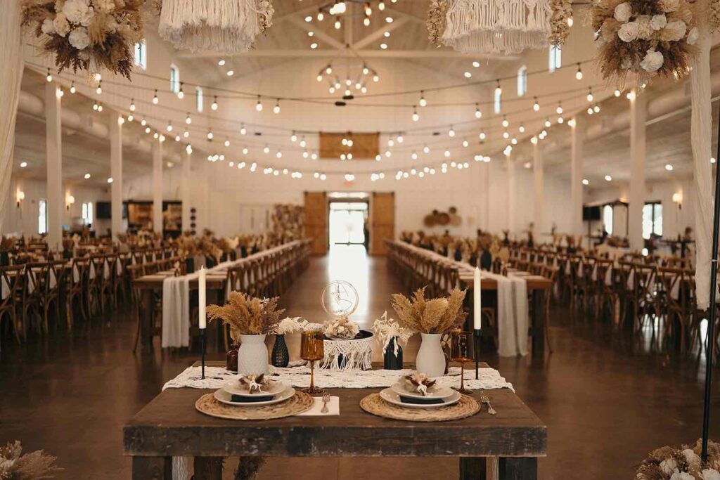 A beautifully decorated wedding reception hall with rustic tables adorned with dried floral arrangements and amber glass vases. Chandeliers with macrame and dried florals hang from the ceiling amidst string lights for a romantic ambiance.