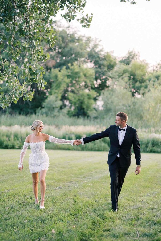 Bride and groom walking hand-in-hand through the lush greenery at The Pines, smiling and enjoying their romantic moment as they stroll through the outdoors. The bride is wearing a stunning off-the-shoulder dress, and the groom looks dashing in his tuxedo.