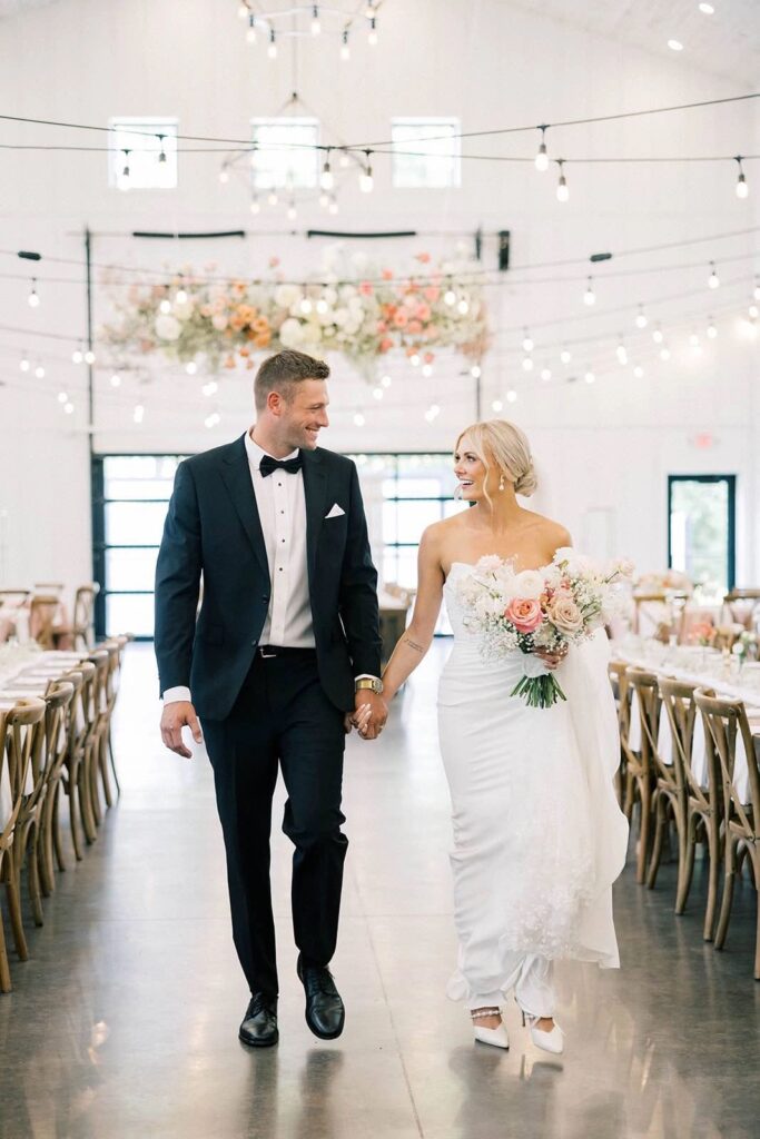 Bride and groom walking hand-in-hand through The Pines reception hall, smiling at each other as they enjoy their first moments together as a married couple. The venue is adorned with soft, romantic floral arrangements and warm string lights overhead.