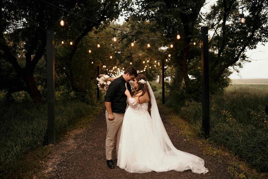 A magical evening portrait of the bride and groom under a canopy of string lights in a wooded path. The groom embraces the bride, who holds a bouquet of dried florals, creating a dreamy atmosphere.