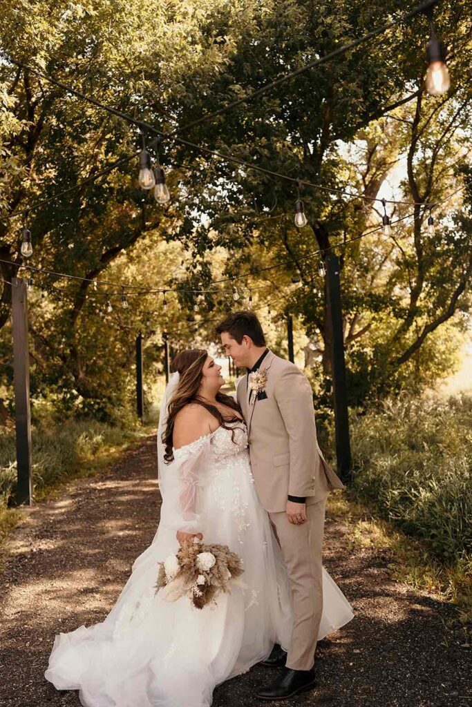 A romantic daytime shot of the bride and groom smiling at each other along a rustic tree-lined path with string lights strung above, adding charm to the natural setting.