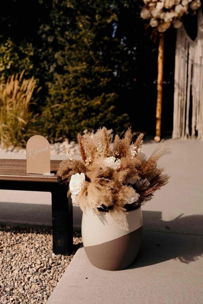 Close-up of a wedding aisle decor featuring a two-tone ceramic vase filled with dried florals, pampas grass, and white roses. A “reserved” sign is placed nearby.