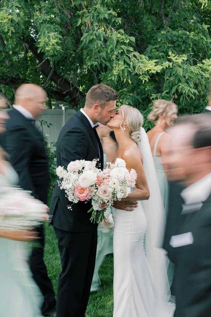 Bride and groom sharing a kiss surrounded by their wedding party, with soft motion blur capturing the joy and movement of the celebration.