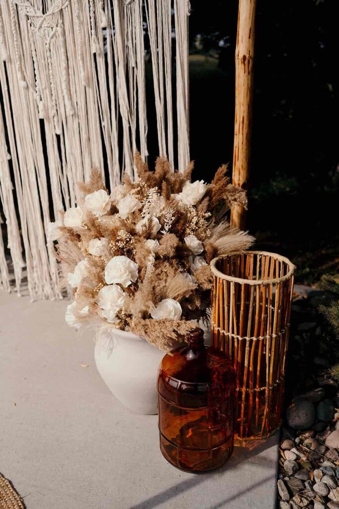 A corner of the wedding ceremony area featuring a white vase with dried floral arrangements, a bamboo lantern, and amber glass accents against a macrame backdrop for a bohemian touch.