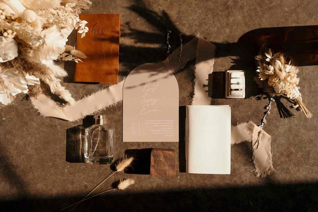 A flat lay composition featuring vow books, a floral boutonniere, perfume, wedding rings, and an invitation, complemented by dried florals and soft fabric ribbons.