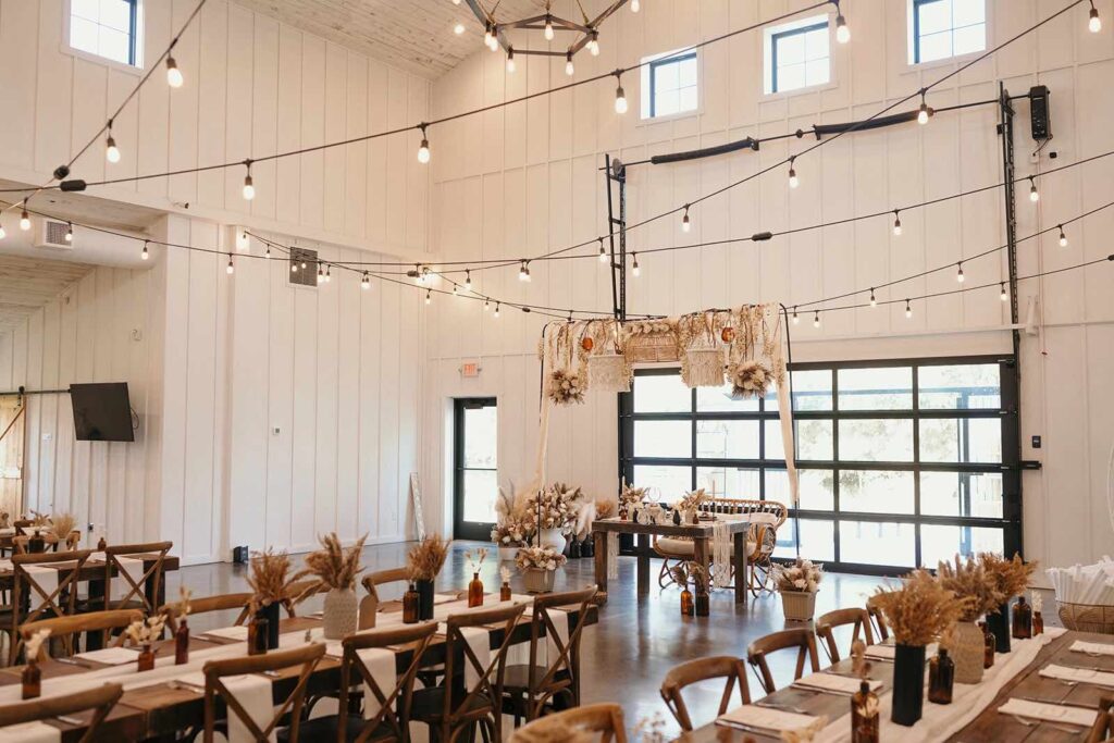 A wide view of a wedding reception setup with long wooden tables decorated with dried floral centerpieces and string lights crisscrossing above, creating an inviting and warm atmosphere.