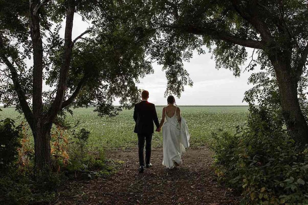 Bride and groom walking hand in hand toward the sunset in a scenic field at The Pines