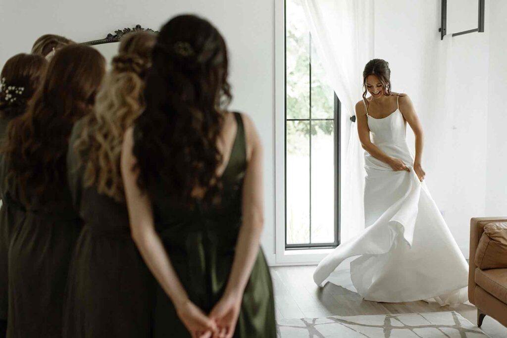 Bride joyfully revealing her wedding dress to her bridesmaids, creating a touching moment