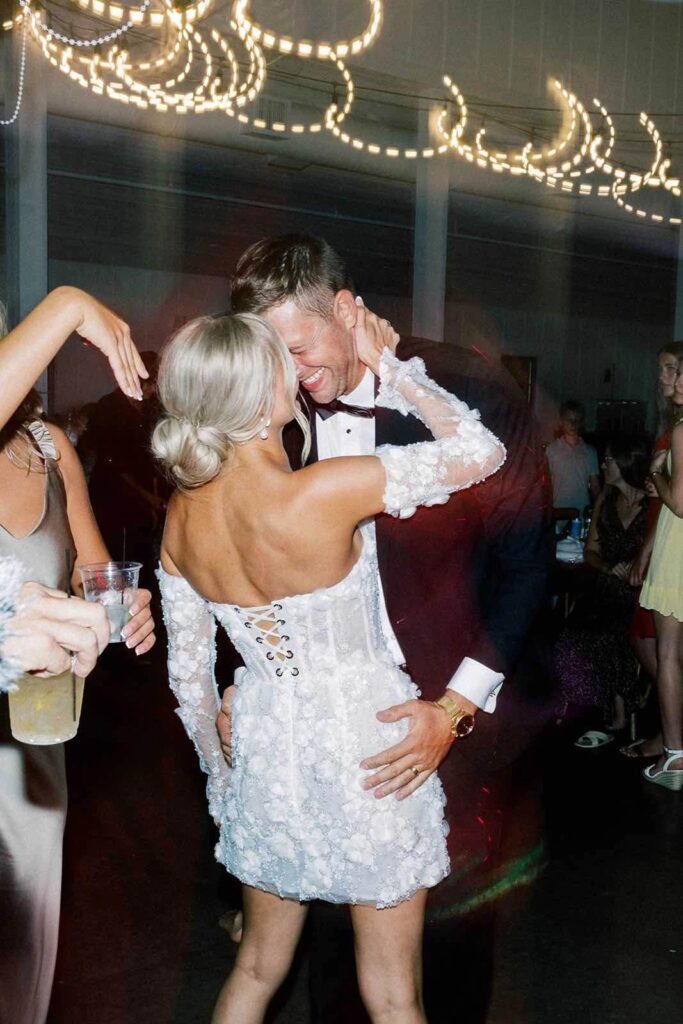 Bride in a stunning off-the-shoulder mini wedding dress dancing with her groom under twinkling reception lights at The Pines.