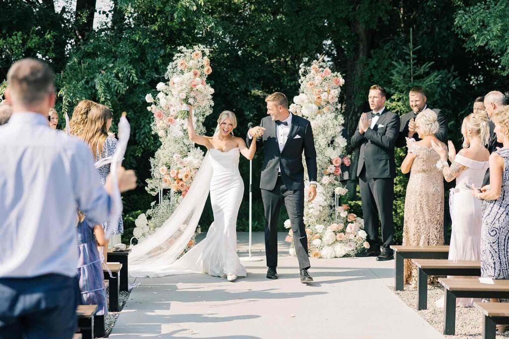 Bride and groom joyfully walking down the aisle after their ceremony at The Pines, holding hands and smiling, surrounded by guests applauding. Stunning floral arrangements of soft pink roses and white flowers frame the outdoor setting.