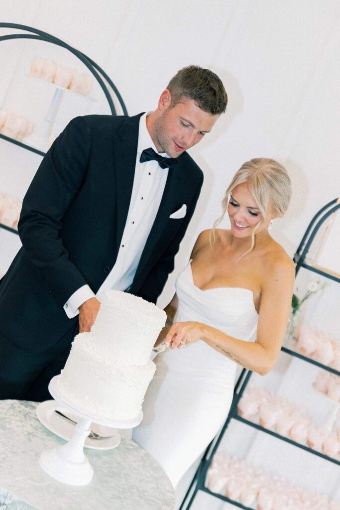 Bride and groom smiling as they cut their wedding cake at The Pines. The couple is surrounded by elegant decor, with a two-tiered white cake adorned with delicate icing, and a backdrop of soft pastel-colored cupcakes.
