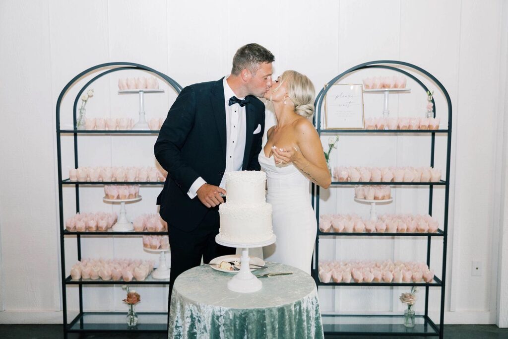 Bride and groom sharing a kiss while cutting their wedding cake at The Pines, surrounded by a display of pink cupcakes and elegant decor. The bride holds a piece of cake as they enjoy their special moment together.