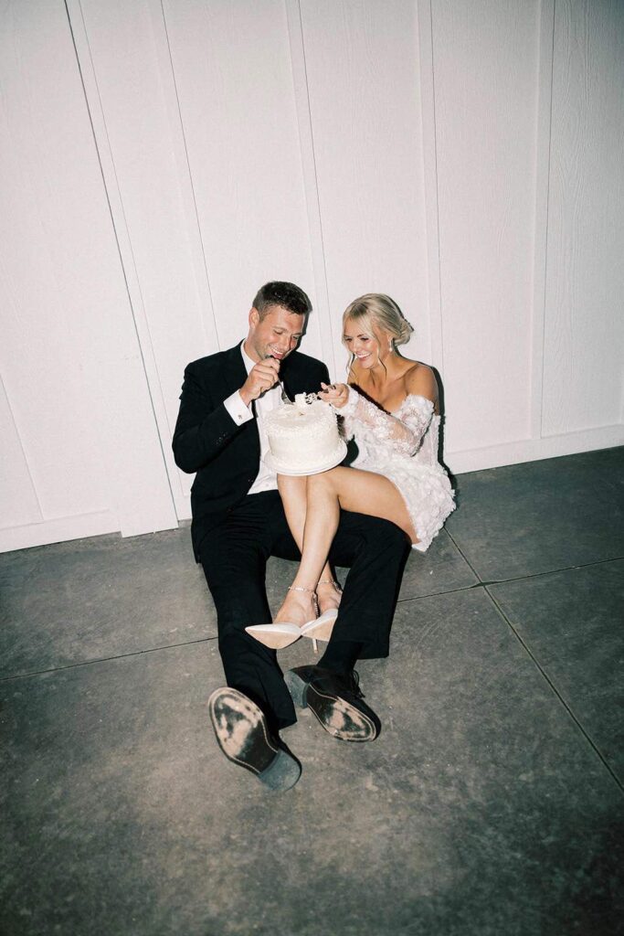 Bride and groom sitting on the floor, laughing and sharing a piece of wedding cake in a fun, intimate post-reception moment.
