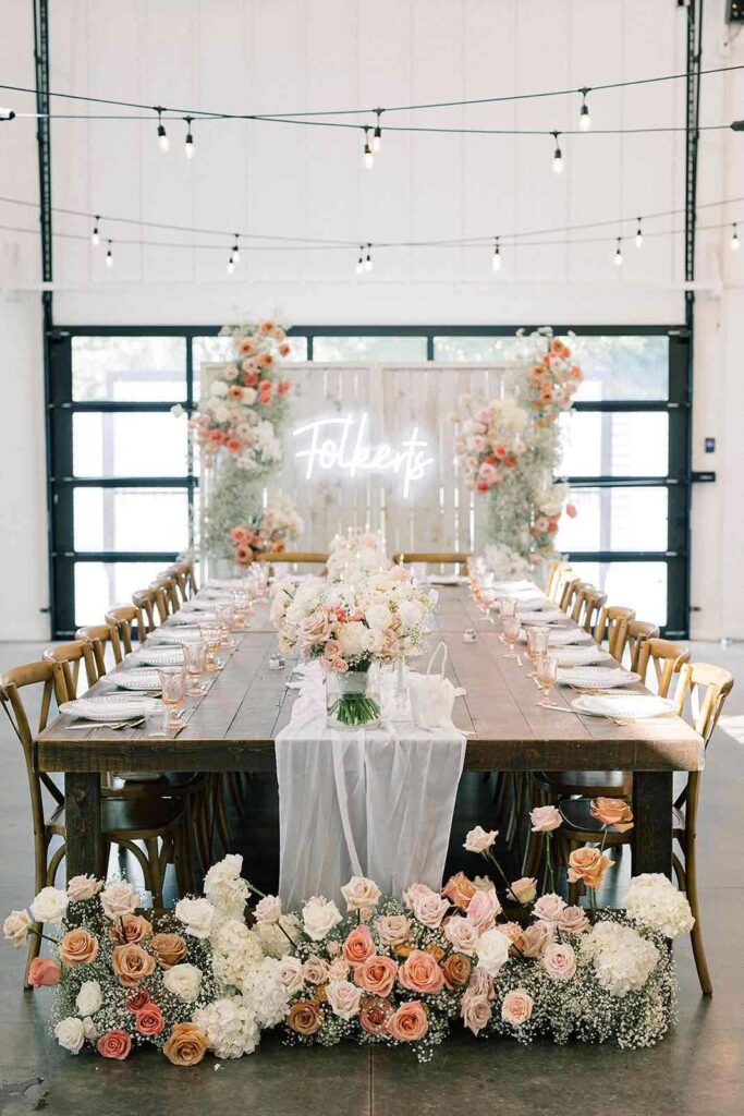 A gorgeously styled wedding reception head table with cascading florals, elegant tableware, and a neon "Folberts" sign in the background.