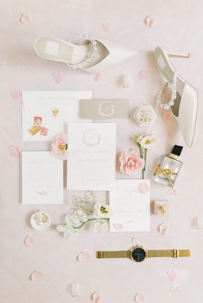 Elegant wedding details flat lay featuring pearl-adorned bridal shoes, invitation suite, rings, perfume, and florals in a soft neutral palette.