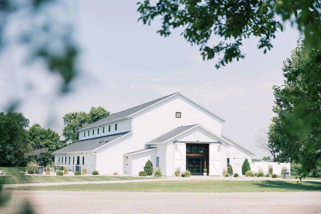 Exterior view of The Pines White Barn wedding venue, showcasing its spacious, modern design with large barn doors, manicured grounds, and a clear blue sky in the background.