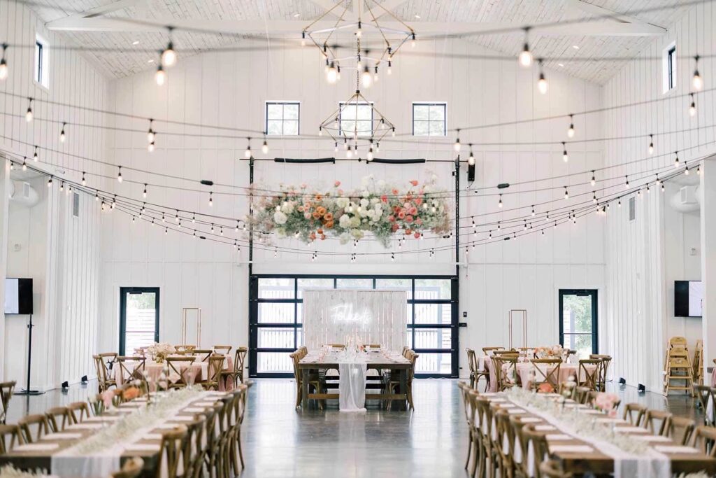 Beautifully designed wedding reception at The Pines White, featuring long farmhouse tables, string lights, and a floral hanging installation.