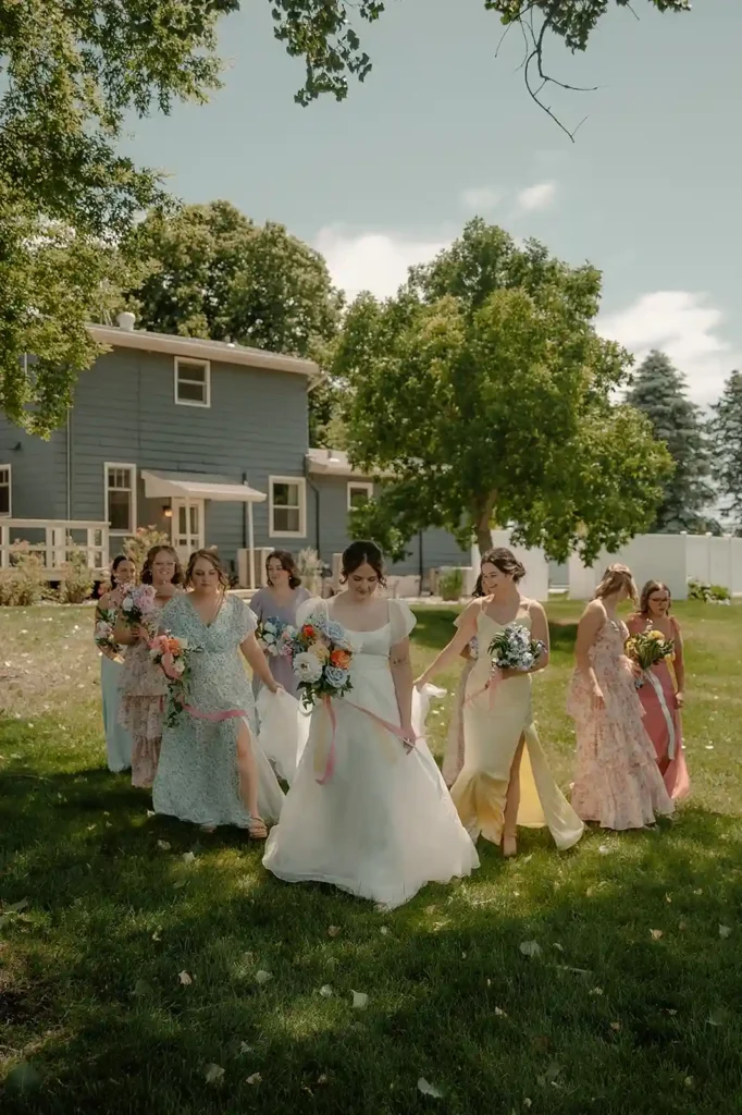 Bride and bridesmaids walk through the lawn wearing colorful summer dresses and holding bright floral bouquets